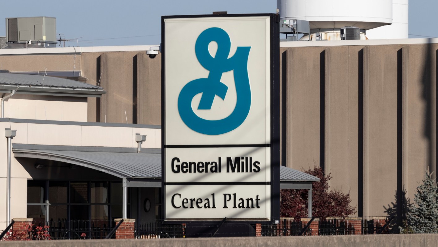 A sign for the General Mills Cereal Plant featuring a blue logo and text in front of a manufacturing facility.