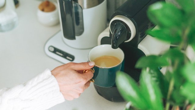 A person pours coffee into a blue mug from a coffee machine on a bright kitchen countertop at home.
