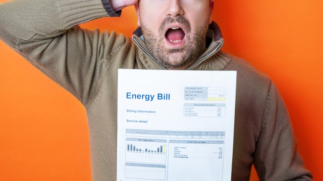 A concerned man holding an energy bill with an orange background, expressing shock with his mouth open and hand on his head.