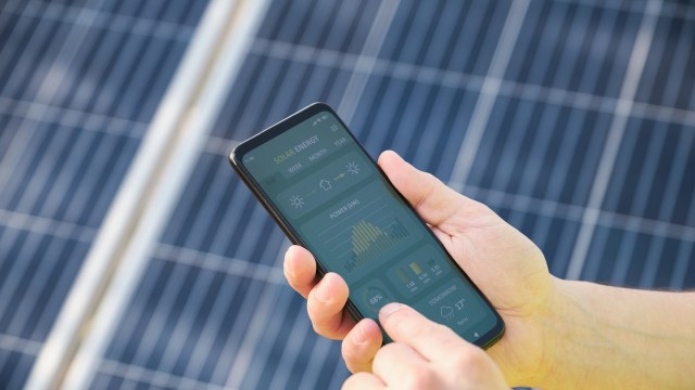 A person uses a smartphone to monitor solar energy data, with solar panels visible in the background.