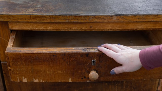 A hand rests on an open wooden drawer of a vintage chest, revealing an empty interior.
