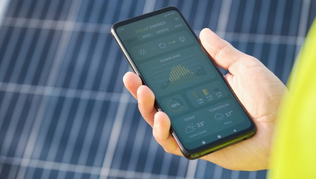 A person in a bright yellow shirt holds a smartphone displaying solar energy data against a backdrop of solar panels.