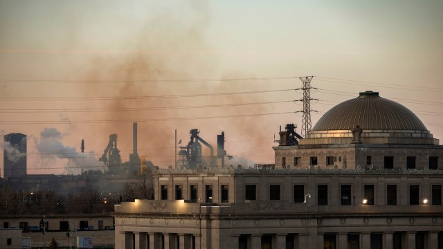 A historic building foregrounded by industrial smokestacks and pollution under a hazy sky.