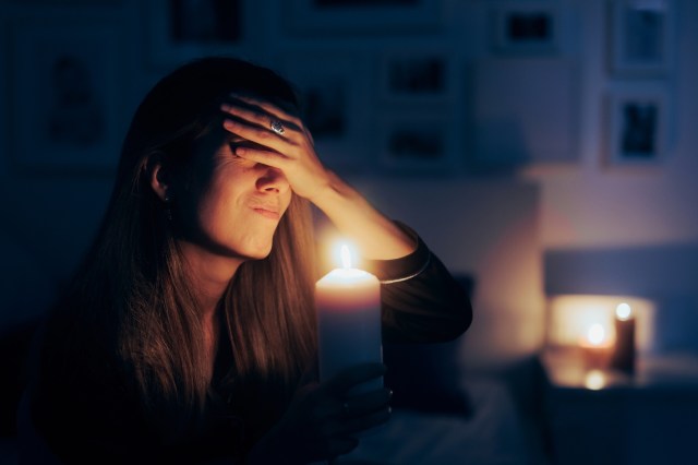 A woman holds a candle while sitting in a dimly lit room with her hand on her forehead.