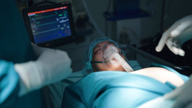 A patient with a breathing mask lies on an operating table, monitored by medical staff in a surgical setting.