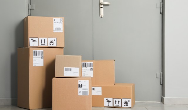 Several stacked cardboard boxes with shipping labels in front of a closed gray door.