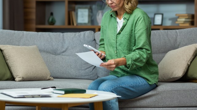A woman with a smartphone reviewing documents in a living room.