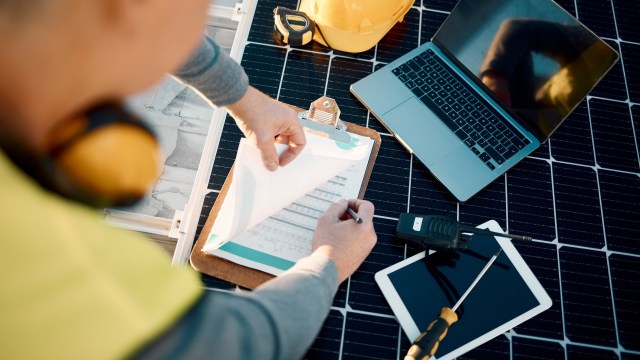 A worker reviews paperwork on a clipboard while using technology on a solar panel installation site.