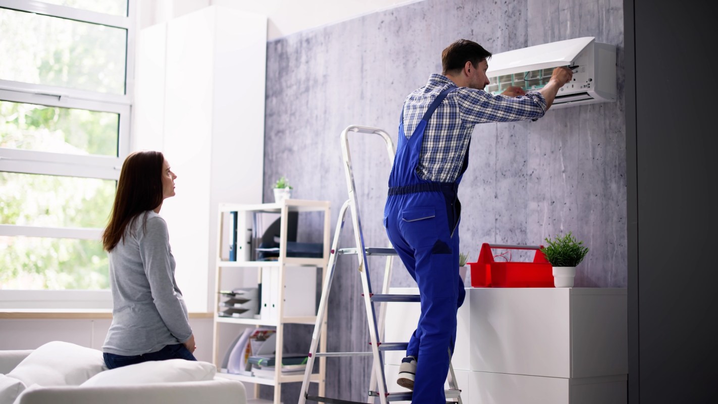 A technician in blue overalls installing an air conditioning unit while someone observes.