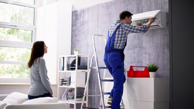 A technician in blue overalls installing an air conditioning unit while someone observes.