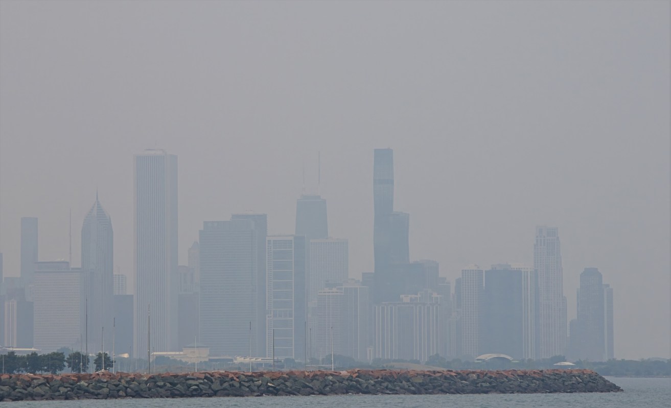 A hazy Chicago skyline of tall buildings peeking through wildfire smoke over a rocky shoreline.