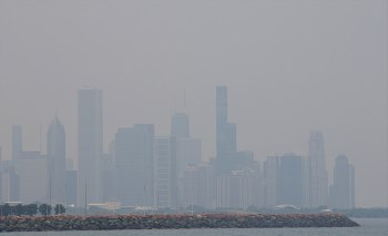 A hazy Chicago skyline of tall buildings peeking through wildfire smoke over a rocky shoreline.