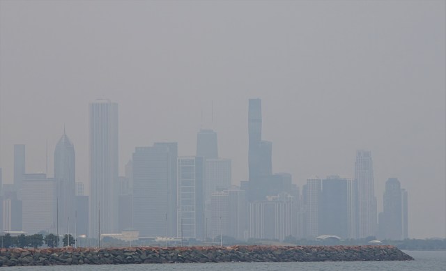 A hazy Chicago skyline of tall buildings peeking through wildfire smoke over a rocky shoreline.