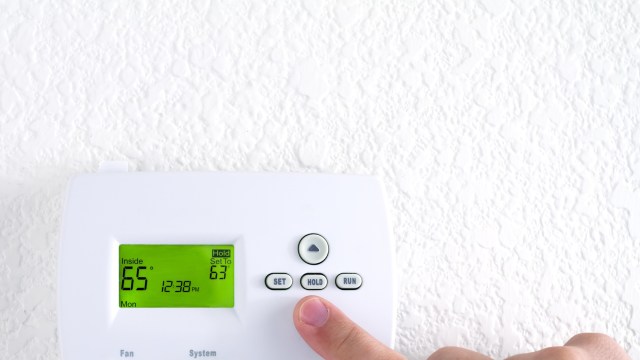 A person adjusting a digital thermostat displaying 65°F.
