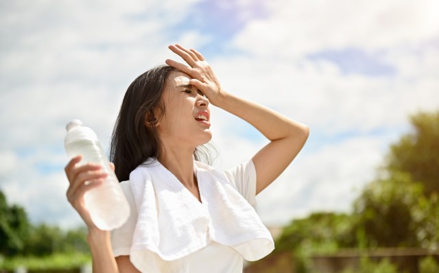 A woman wipes her forehead, holding a water bottle and wearing a towel around her neck, in an outdoor setting.