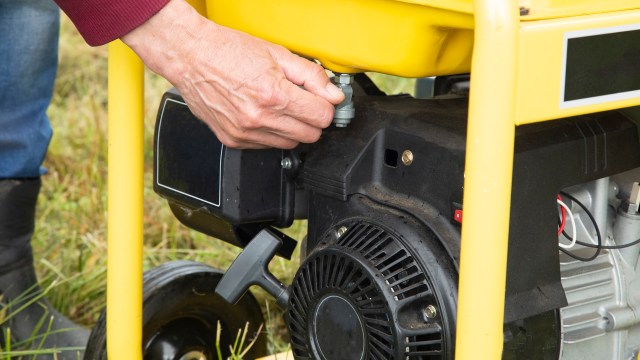 A person is adjusting the fuel valve on a yellow small engine in a grassy outdoor setting.