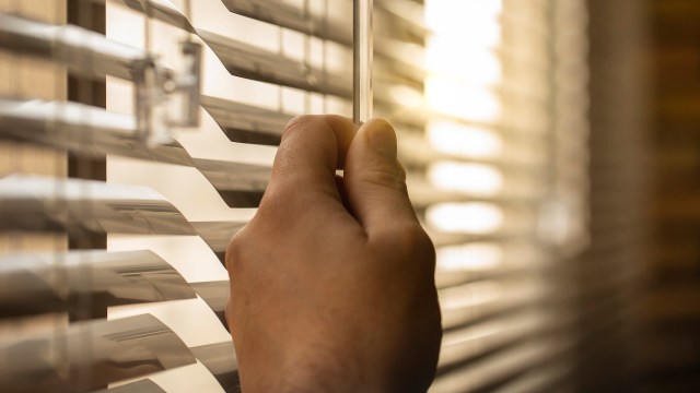 A hand adjusting horizontal blinds with warm sunlight illuminating the scene.