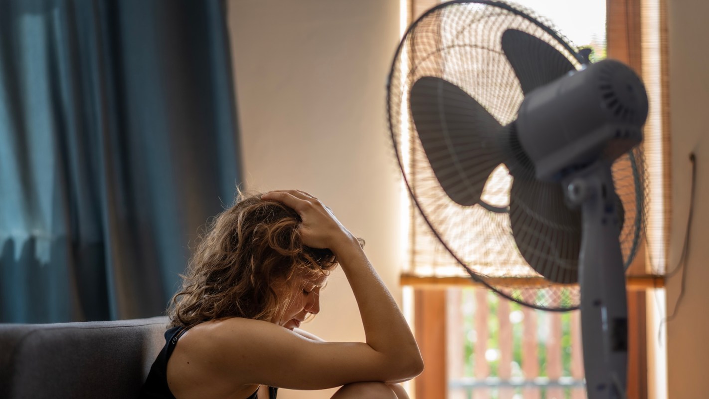 A woman sitting on a couch with her head resting on her knees, looking stressed near an electric fan.