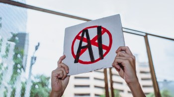 A person holds a handmade sign with a red circle and a diagonal line over the letters "AI."