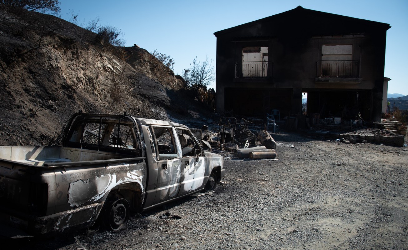 A burnt-out truck and a blackened house stand amid charred earth and debris in a scorched landscape.