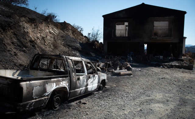 A burnt-out truck and a blackened house stand amid charred earth and debris in a scorched landscape.