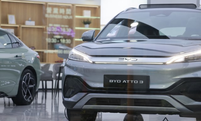 A grey BYD Atto 3 electric vehicle displayed in a showroom with another car partially visible in the background.