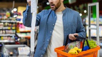 A man holds a long receipt while examining his grocery basket filled with fruits and vegetables in a store.