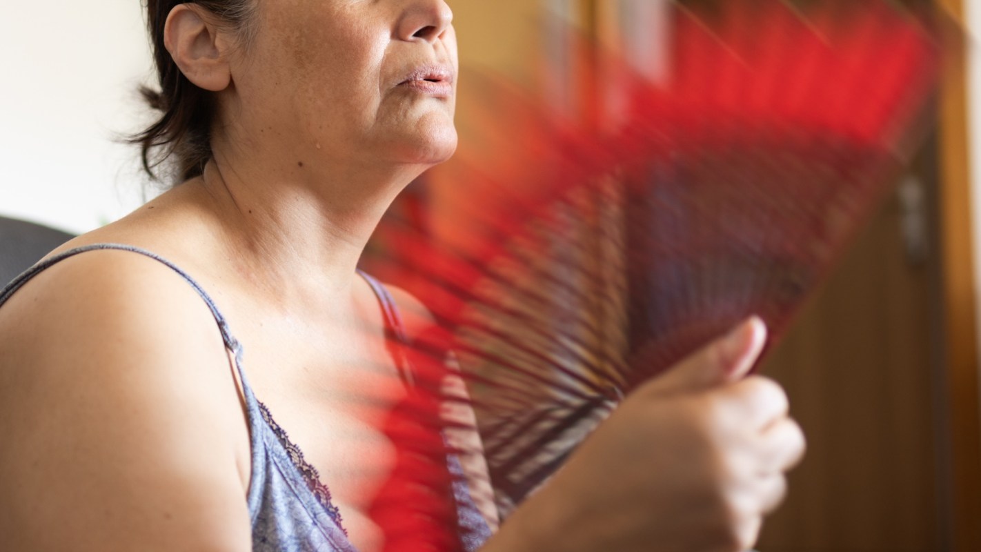 A woman fans herself with a red fan while looking thoughtfully, with a blurred background of a room.