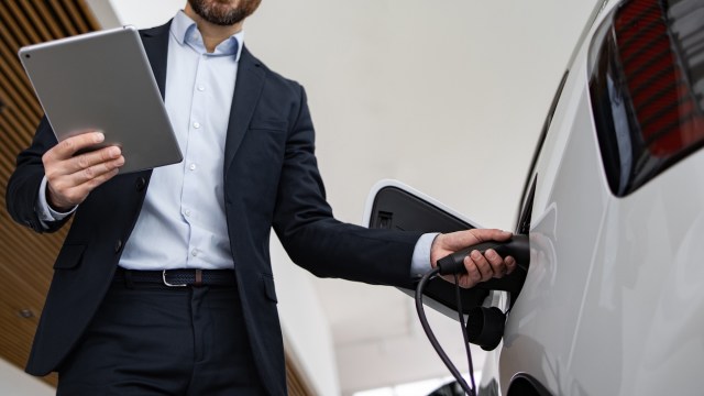 A man holding a tablet while plugging a charger into an electric vehicle.