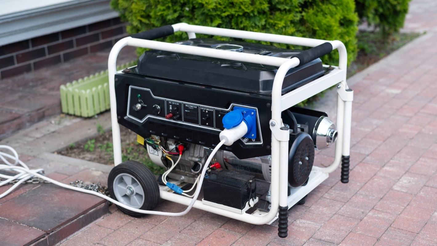 A portable generator with wheels, power connections, and control panel on a brick pavement near greenery.