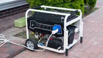 A portable generator with wheels, power connections, and control panel on a brick pavement near greenery.