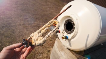 A person holds a corroded heating element with mineral buildup next to a white water heater tank.
