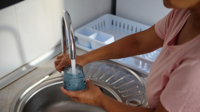 A woman fills a glass with water from a kitchen faucet.