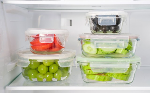 Glass containers with assorted fruits and vegetables, including tomatoes, cucumbers, and grapes, stored in a refrigerator.