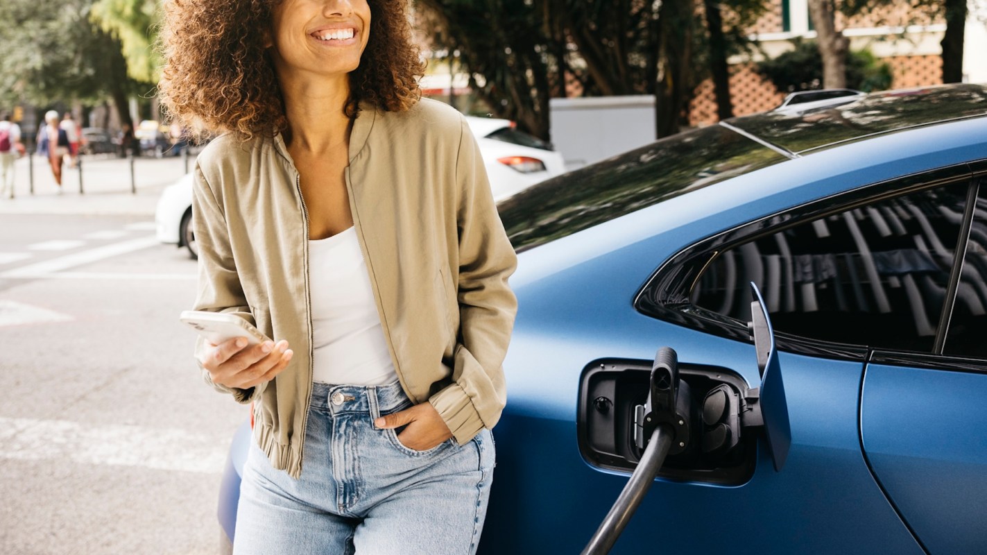A woman stands beside a blue electric car, holding a phone while charging the vehicle.