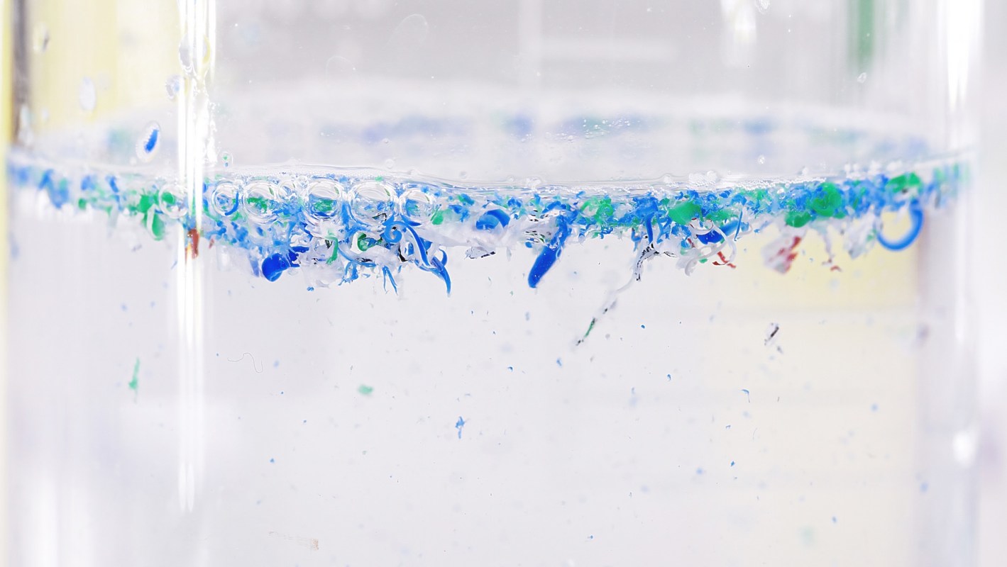 A close-up of a clear container filled with water, showing colorful microplastics suspended near the surface.