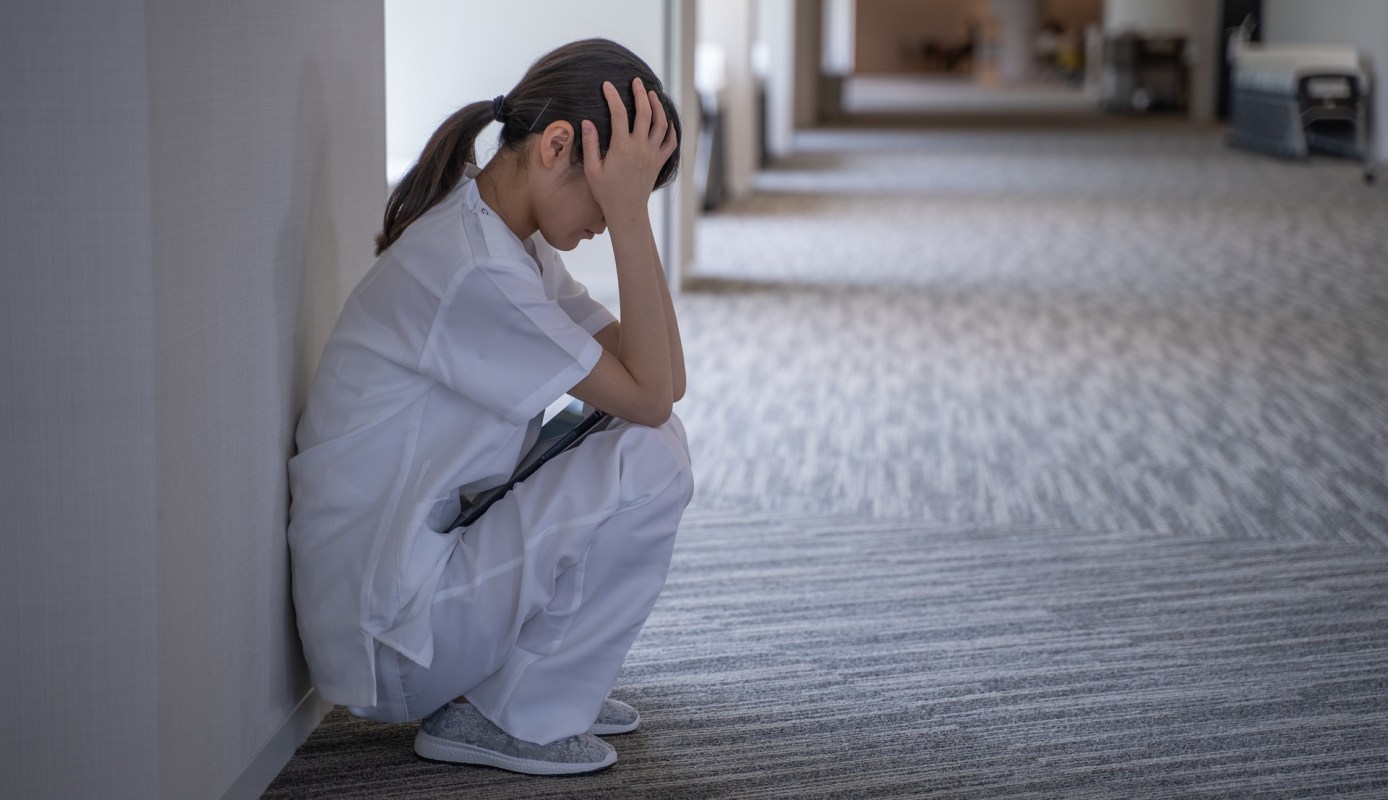 A healthcare worker sits on the floor, holding their head in distress in a hospital corridor.