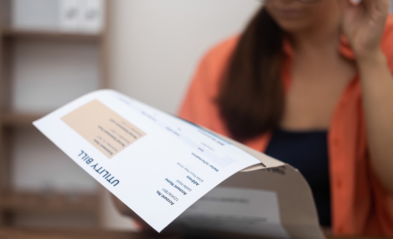 A woman in an orange shirt examines a utility bill while adjusting her glasses.