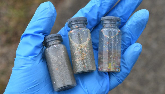 A hand in a blue glove holds three small glass vials containing colorful sediment and particles.