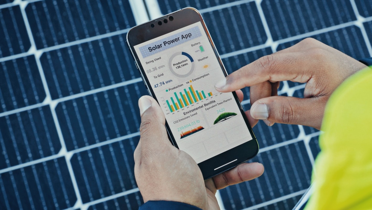A person uses a smartphone app to monitor solar power production and environmental benefits with solar panels in the background.