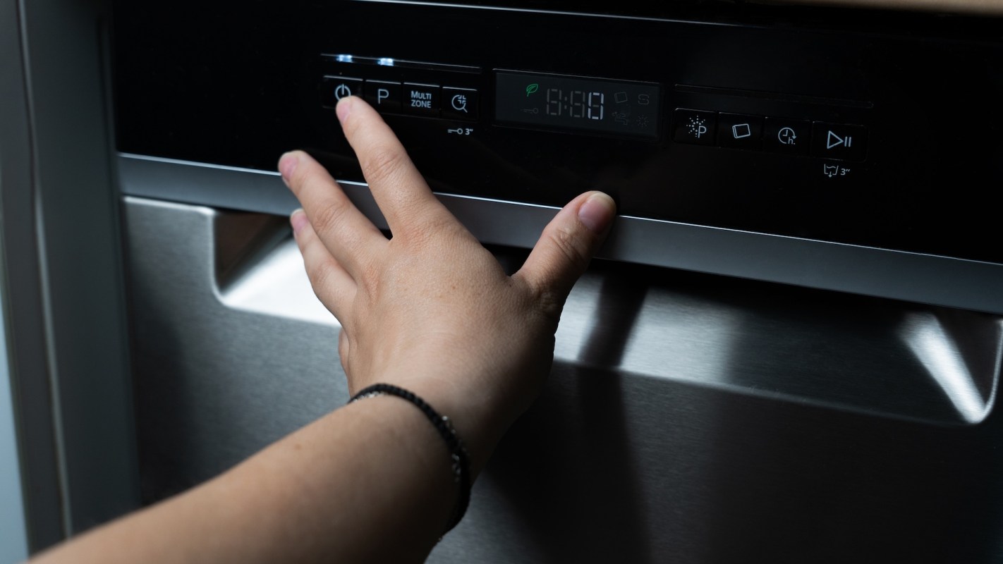 A hand is adjusting the control panel of a dishwasher in a dark kitchen.