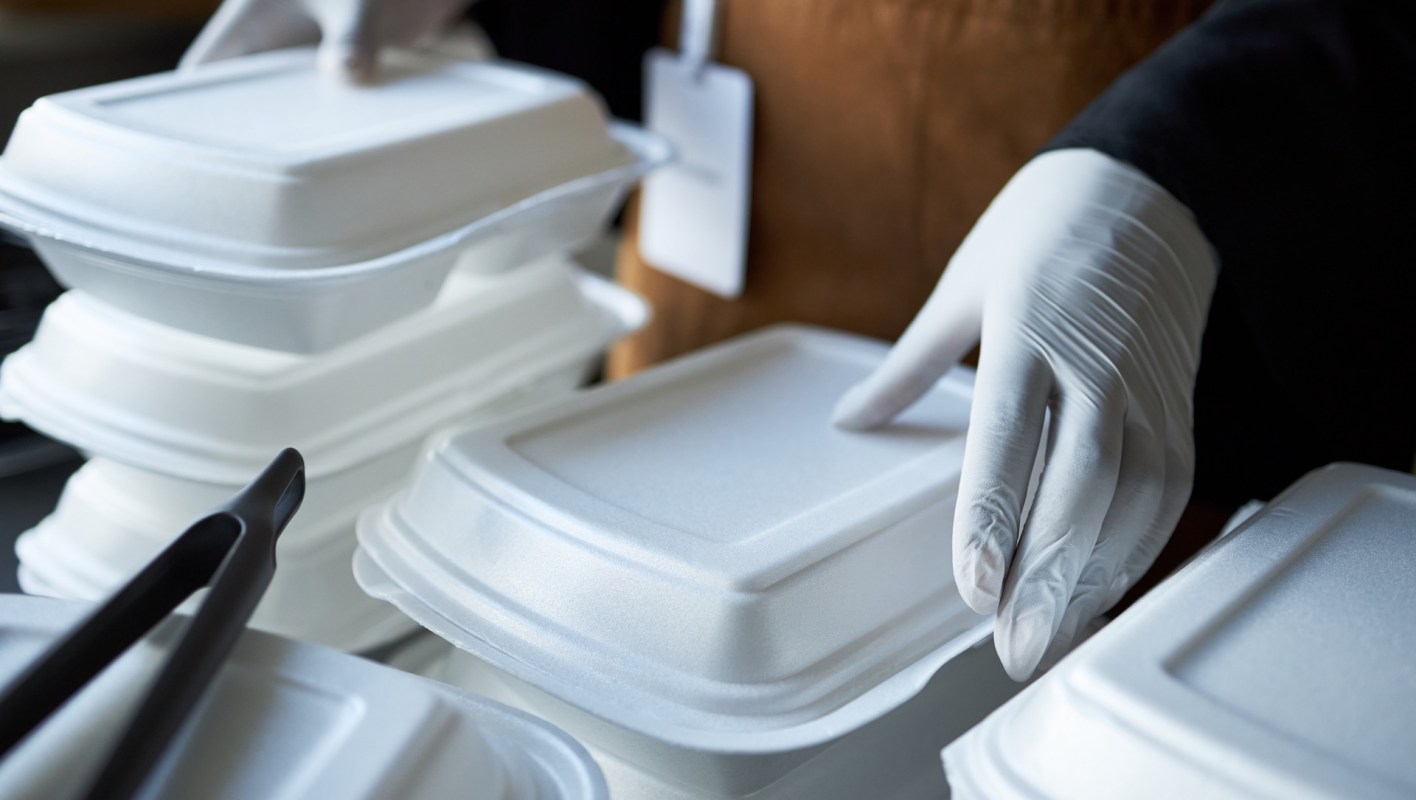A person in gloves is carefully handling Styrofoam takeout containers stacked on a surface.