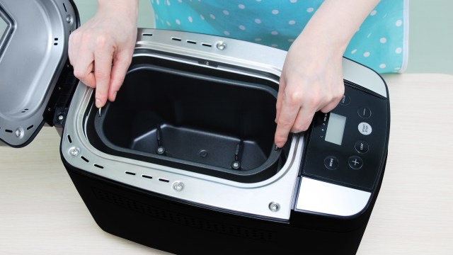 A person adjusts the interior of a bread maker while its lid is open on a light wooden surface.