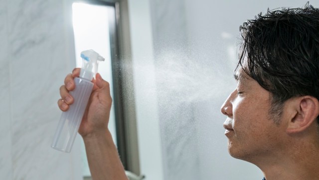 A man sprays mist from a bottle onto his face while standing in a bright bathroom.