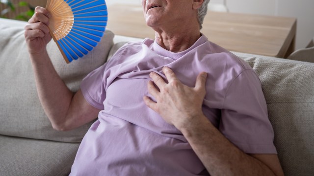 An elderly man wearing a purple shirt fans himself while sitting on a couch, putting his hand over his heart.