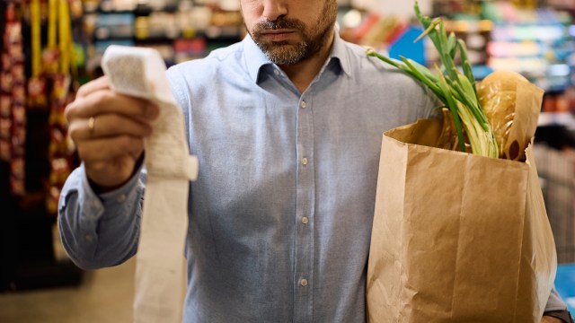 A man in a blue shirt examines a grocery receipt while holding a paper bag with vegetables.