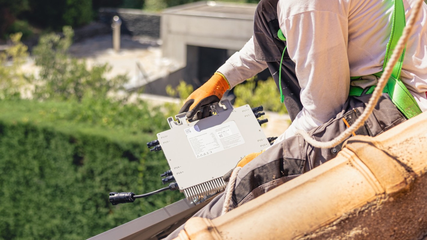 A worker in gloves holds a solar inverter while perched on a rooftop, overlooking greenery and buildings below.
