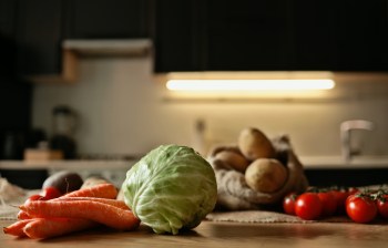 A variety of fresh vegetables including cabbage, carrots, tomatoes, and potatoes on a wooden countertop in a dark kitchen.