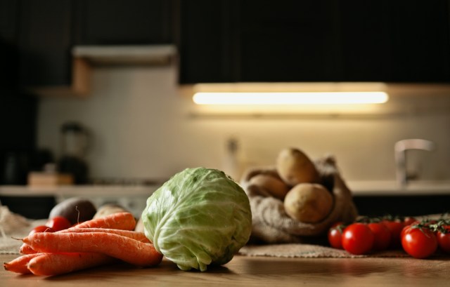 A variety of fresh vegetables including cabbage, carrots, tomatoes, and potatoes on a wooden countertop in a dark kitchen.