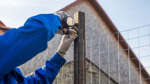 A person in gloves secures a rusty metal fence post with a drill against a wooden structure in the background.
